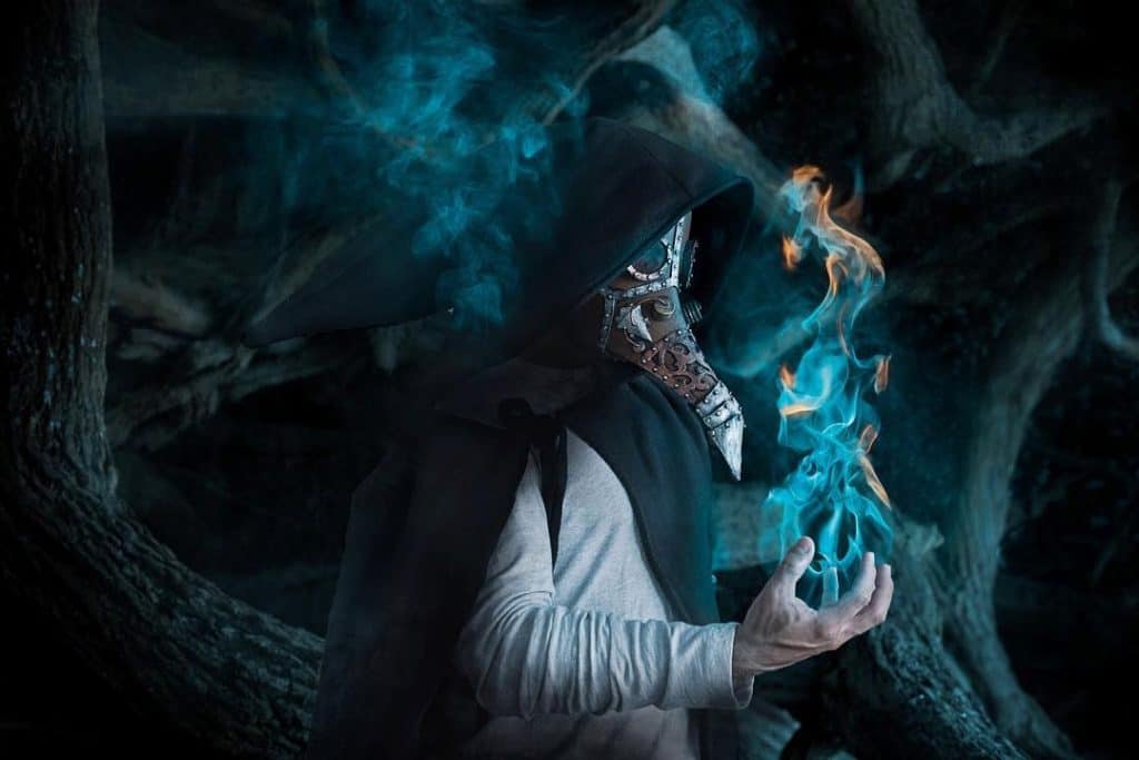 A fine art composite photo with a person wearing a black cape and plague doctor mask. He is holding blue and orange flames in his right hand.