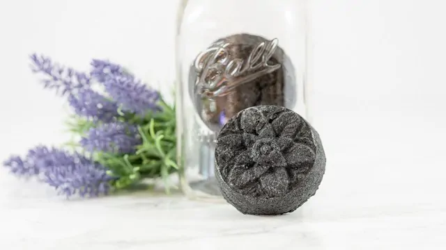 A black bath bomb made from activated charcoal leaning against a glass mason jar containing two additional black bath bombs. Lavender is visible in the background