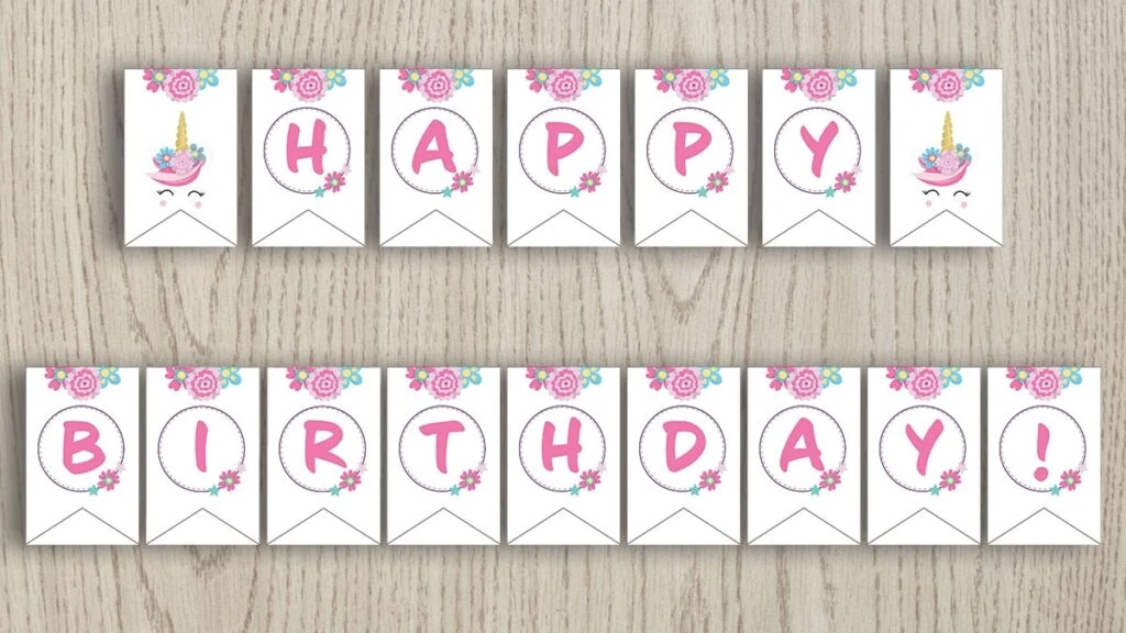 Free printable happy birthday banner with pink flowers and unicorns. The banner is shown on a wood background. 