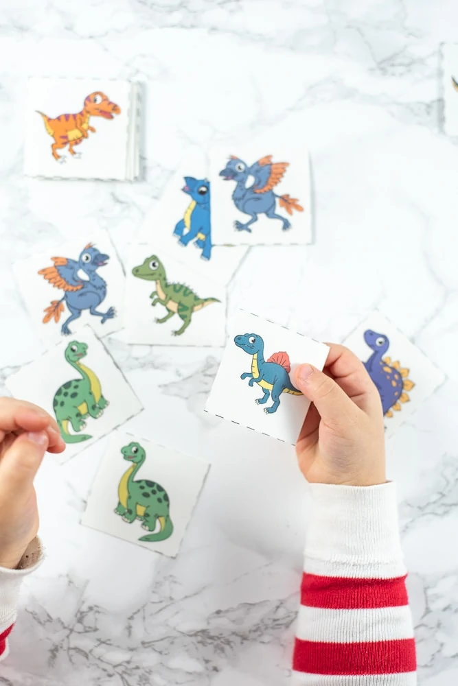A toddler playing with a matching game featuring mirror image pairs of cartoon dinosaurs. She is holding a green brachiosaurus and a spinosaurus in her hands. Only her hands and arms, in red and white striped pajamas, are visible, not the rest of her body. In the background there are more dinosaur matching cards on a white faux marble surface.