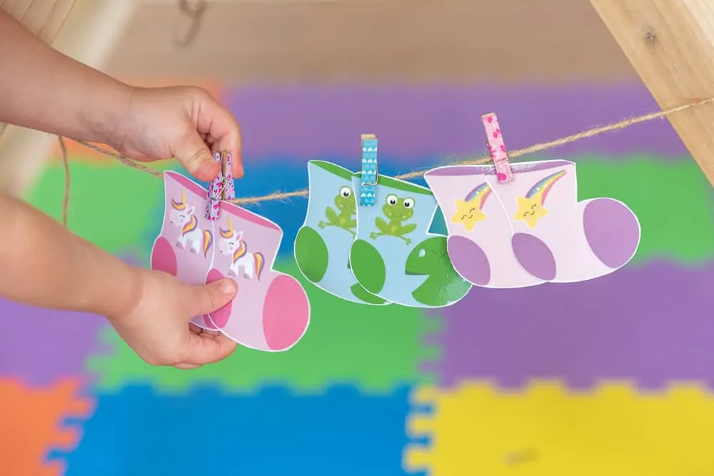 a toddler unclipping a pair of printable unicorn socks from a piece of twine. The socks are hanging on a limeade from twine with two more pairs of printable socks from a printable sock matching game. The other pairs of socks are green with frogs and pink with stars, respectively. The toddler's hands and arms are in frame.
