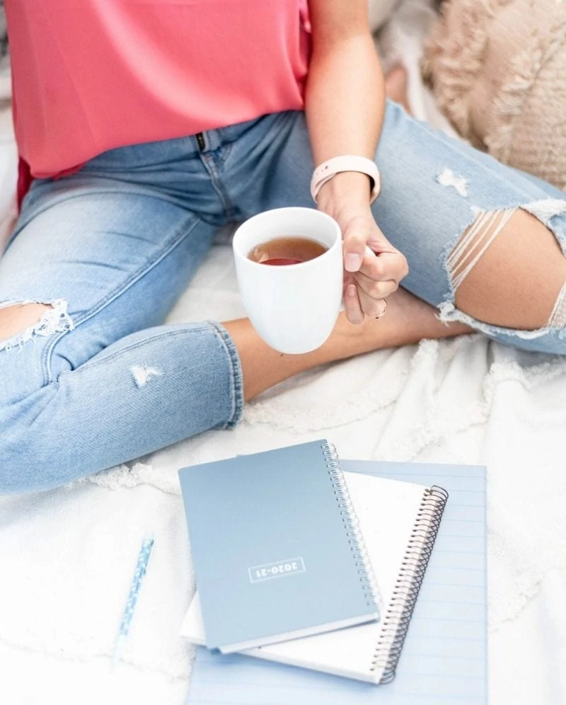 A woman with ripped knee jeans sitting on a white blanket with blue spiral notebooks and a mug of tea