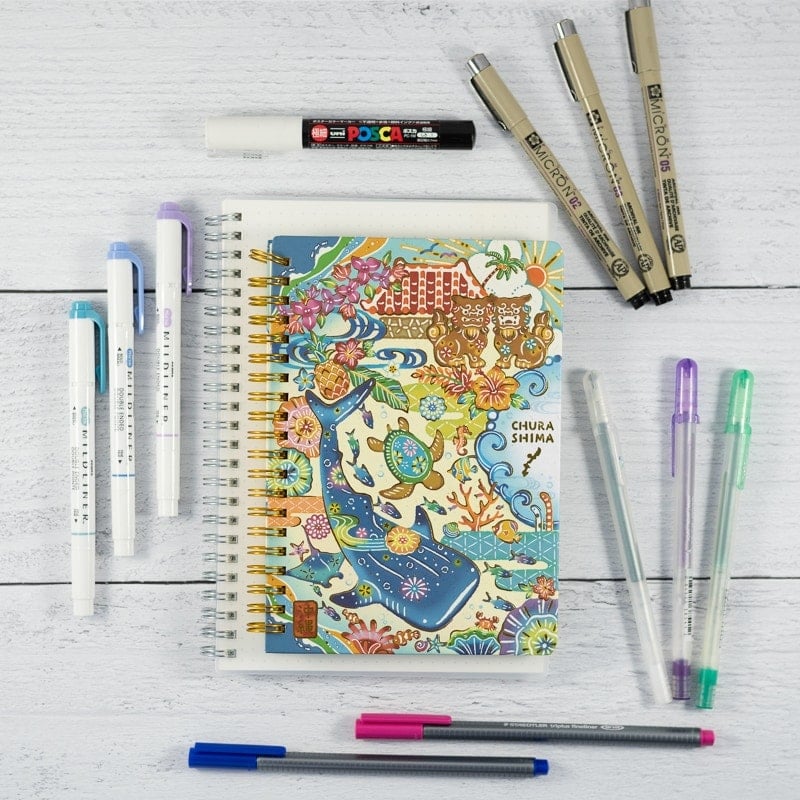 An image with a spiral Japanese notebook and a collection of bullet journal pens including Sakura Microns, gelly roll pens, Zebra midliners, and fineliner markers