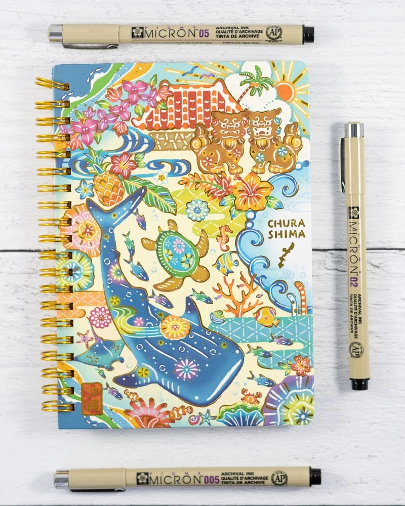 Micron pens next to a whale shark themed small spiral notebook