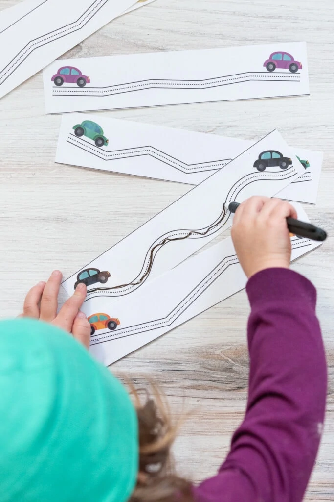 A top down view of a preschooler using a dry erase marker to color in a trace along the lines prewriting practice page with cars on it.