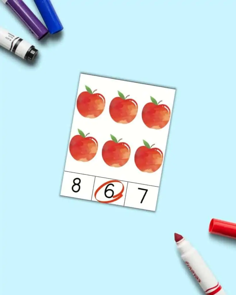 A mockup of a count and clip card with 6 red apples. Below the apples are the numbers 8 - 6 - 7. The 6 is circled in red.