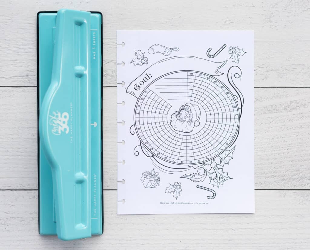 A photo of a printable Santa themed habit tracker. The page has been trimmed to fit a Happy Planner Classic and is shown next to a blue MAMBI Happy Planner hole punch.