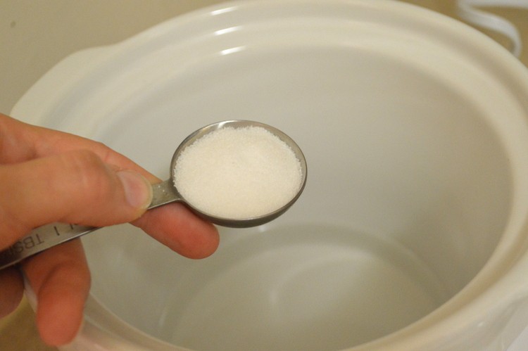 a hand holding a tablespoon of salt over a white crock