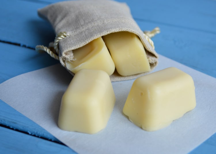 small DIY lotion bars that are shaped like ice cubes. They are in front of a small muslin bag on a  blue wood surface