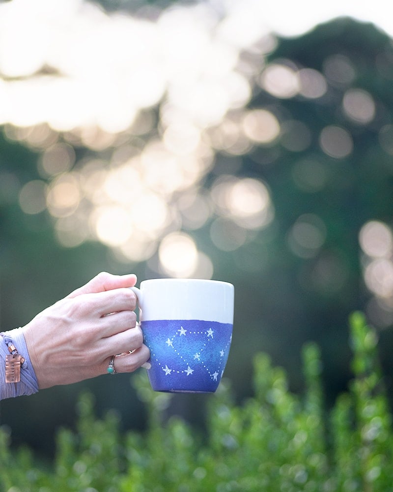 A hand holding a galaxy mug with purple and blue galaxy paint and white stars. 