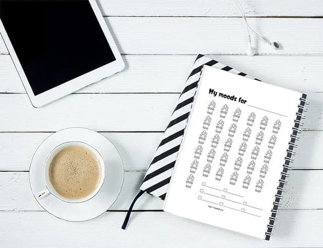 A cactus mood tracker printable in a spiral notebook. It is next to a cup of coffee and a tablet on a white wood table.