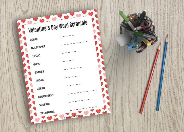 Valentine's Day word scramble with hearts on it on wood next to two pencils and a pen cup
