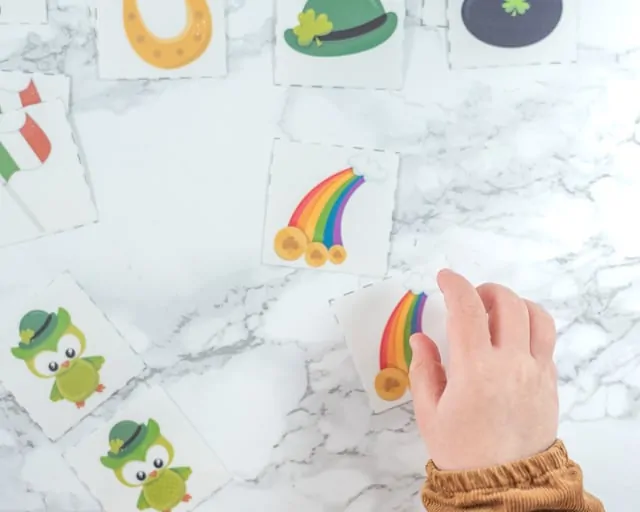 a toddler's hand reaching for a matching card with a rainbow on it. A set of St. Patrick's Day matching cards for toddlers is in the background on a white marble backdrop.