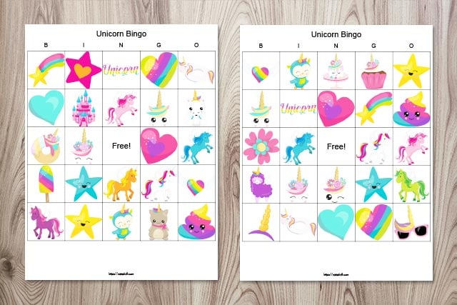 Two free printable unicorn bingo boards on a wood background. The boards feature brightly colored cartoon images with unicorns, hearts, rainbows, and stars.