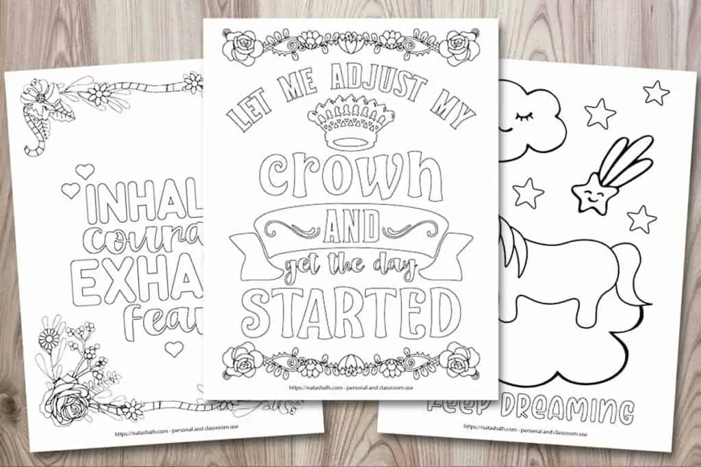 A mockup of three inspirational quote coloring pages on a wood background. The center page has a crown and the text "let me adjust my crown and get the day started." Behind it and to the left is a coloring page with a floral border and the text "inhale courage, exhale fear." On the right is a coloring page with a cartoon unicorn standing on a cloud and the text "keep dreaming"