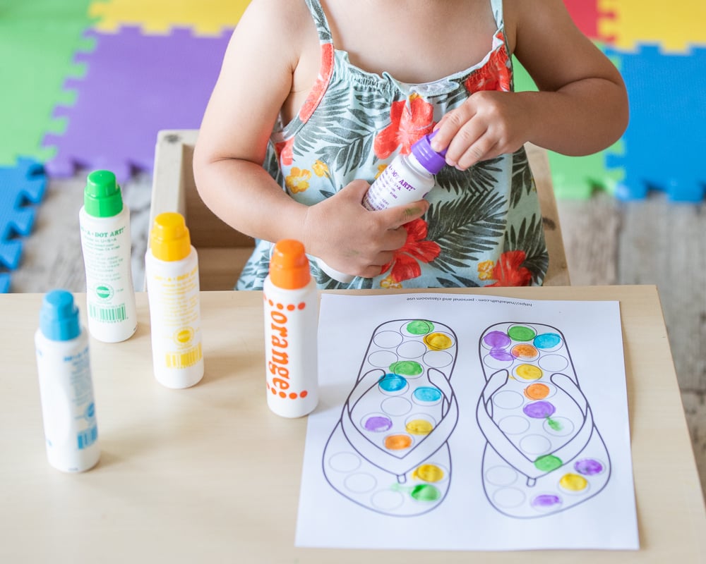 A toddler unscrewing the lid of a purple do a dot marker. Four more markers are visible on a table in front of her. Only the toddlers hands and body are visible.