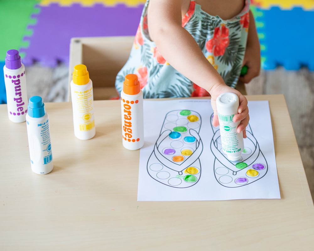 A toddler using a summer themed printable do a dot worksheet showing a sand bucket. The toddler is sitting at a table with five do a dot markers and holding the purple marker. Only the toddler's body and hands are visible. Her face is out of the frame.