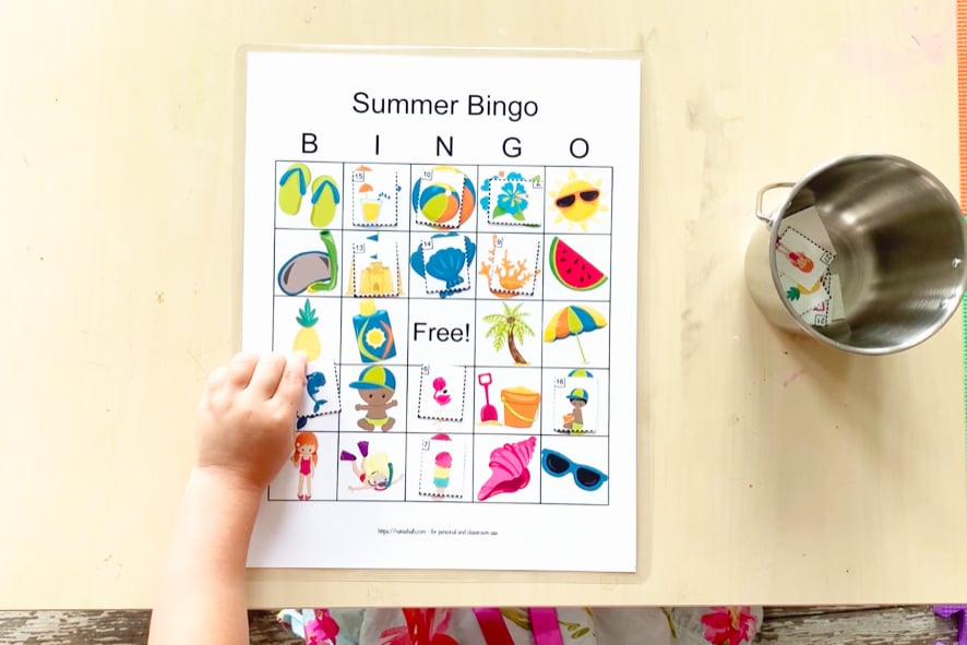 A top down shot of a toddler table with a summer picture bingo card. The toddler is matching a dolphin tile to the dolphin image on her card. Several other pictures are covered and the rest of the call cards are in a metal bucket on the table.