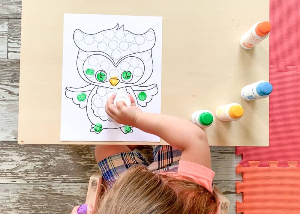 A toddler using colorful dot markers to color a dab a dot marker printable featuring a cartoon owl. The toddler is working at a small table. Colorful foam tiles are visible on the floor. The child's face is not visible because the photo is taken from overhead.