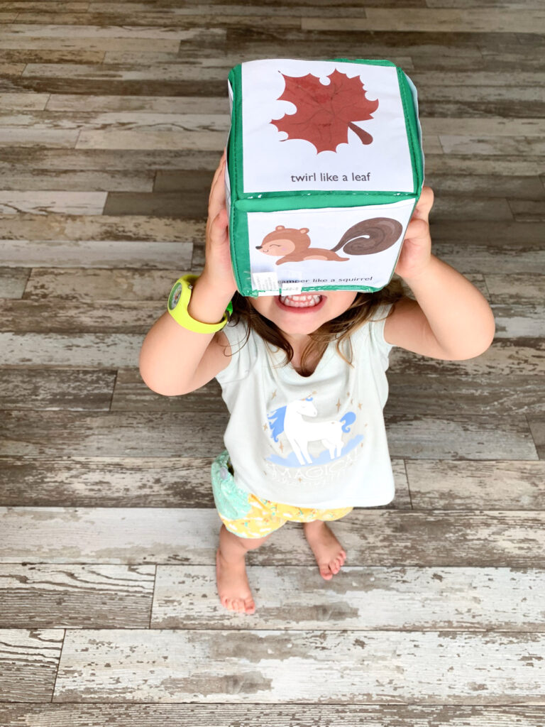 A toddler holding up a differentiated instruction cube with fall themed gross motor movement cards. The top card says "twirl like a leaf". The toddler is wearing a unicorn shirt and a green watch.