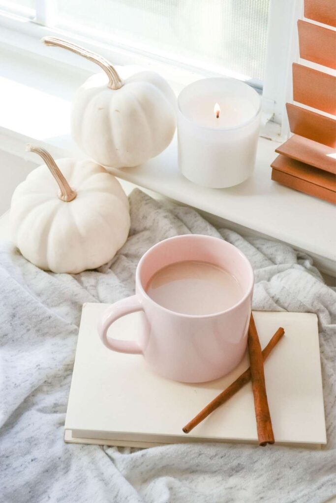a shot of a cozy journaling scene with two white pumpkins, a lit candle on a windowsill, and a pink mug with hot chocolate resting on a white notebook