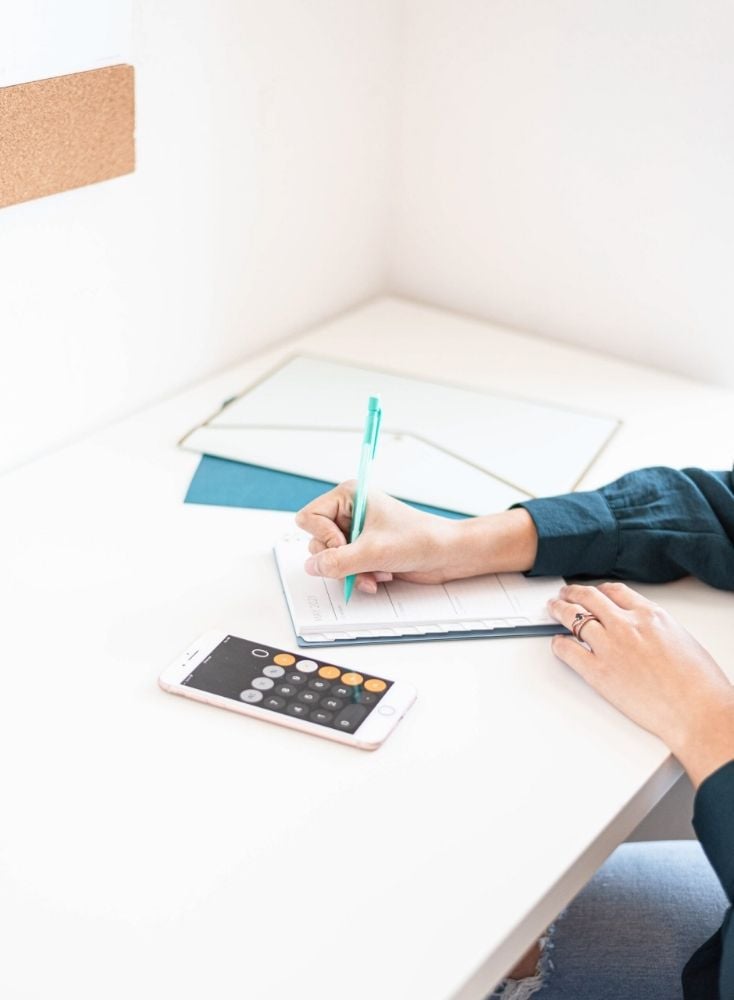 A shot of a woman's hands writing in a planner while adding doing her monthly budgeting. She is holding a green pen and using her iPhone calculator. 