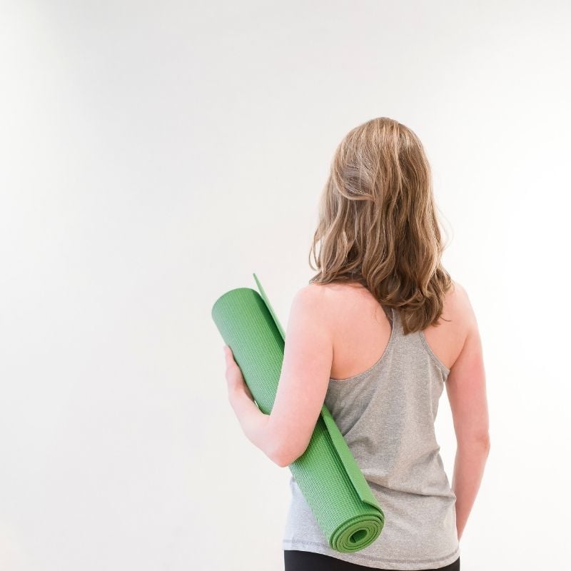 A shot of a woman from the waist up in a grey tank top holding a green yoga mat. She had shoulder length dirty blond hair and is facing a white wall