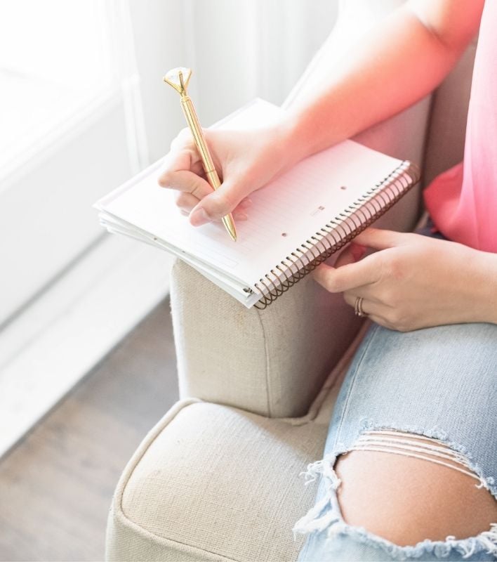 A close up picture of a woman journaling while sitting on a sofa. The woman's hands, knee, and part of her pink shirt are visible. She's holding a large gold pen and writing in a spiral notebook while leaning it on the arm of an oatmeal colored couch.