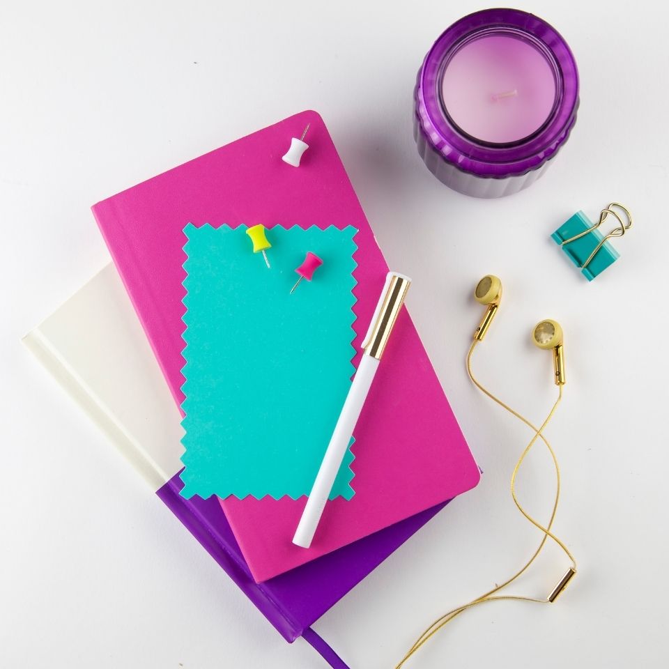 Stationary supplies on a white background. A white and purple notebook is in the middle with a pink notebook and teal notecard on top. A purple candle is above and to the right. A pair of gold earbuds is next to the notebooks.