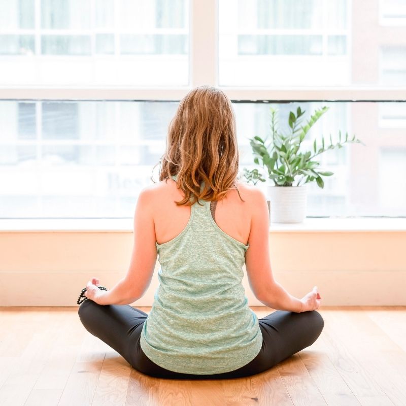 A picture of a woman sitting on a wood floor looking out a window in meditation. She is facing away from the camera and is wearing a green tank top and black yoga pants