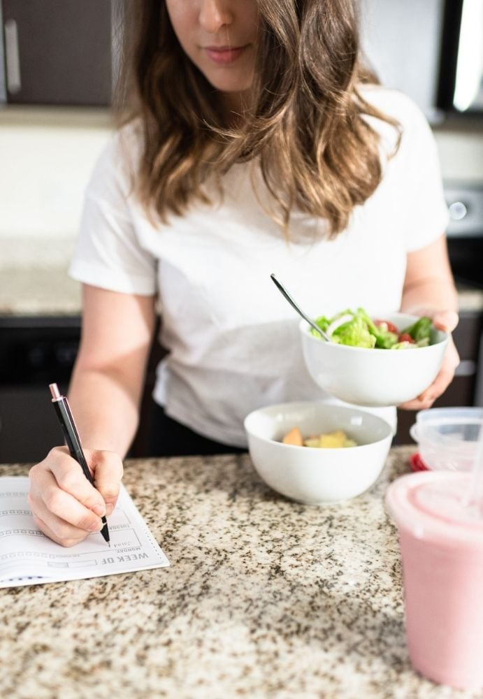 An image of a woman writing in a weekly planner on her kitchen counter while holding a bowl of salad. 