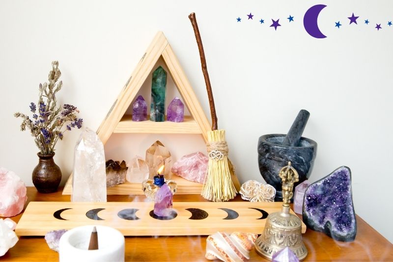 A home altar for celebrating a full moon ritual. There are candles, crystals, a bell, a shell, a burning incense cone, and a vase with dried flowers on a wood table. A triangular shelf with crystals is against the wall with a small broom.
