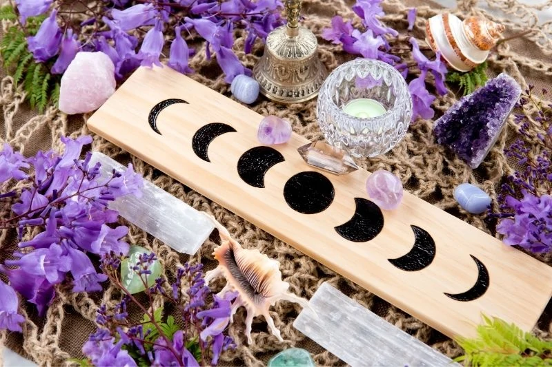 A board with the phases of the moon surrounded by purple bellflowers, two selenite wands, a candle, and amethyst crystals. 