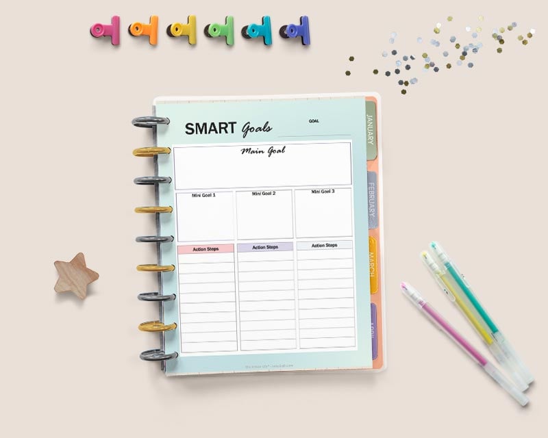 An open Happy Planner with a page reading "smart goals" with spaces to break each goal into three parts with action steps. There are gel pens, glitter, a wood star, and colorful binder clips around the planner.