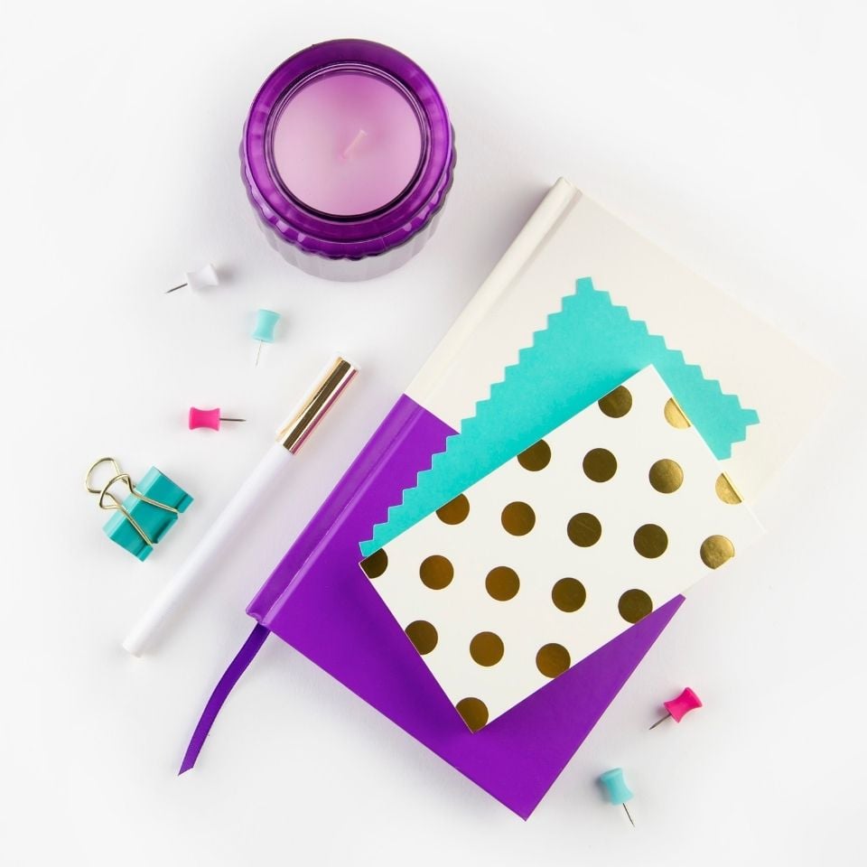 Stationary supplies on a white background. A white and purple notebook is in the middle with teal and gold notecards on top. A purple candle is above and to the left. A teal binder clip, pen, and several push pins are around the notebook. 