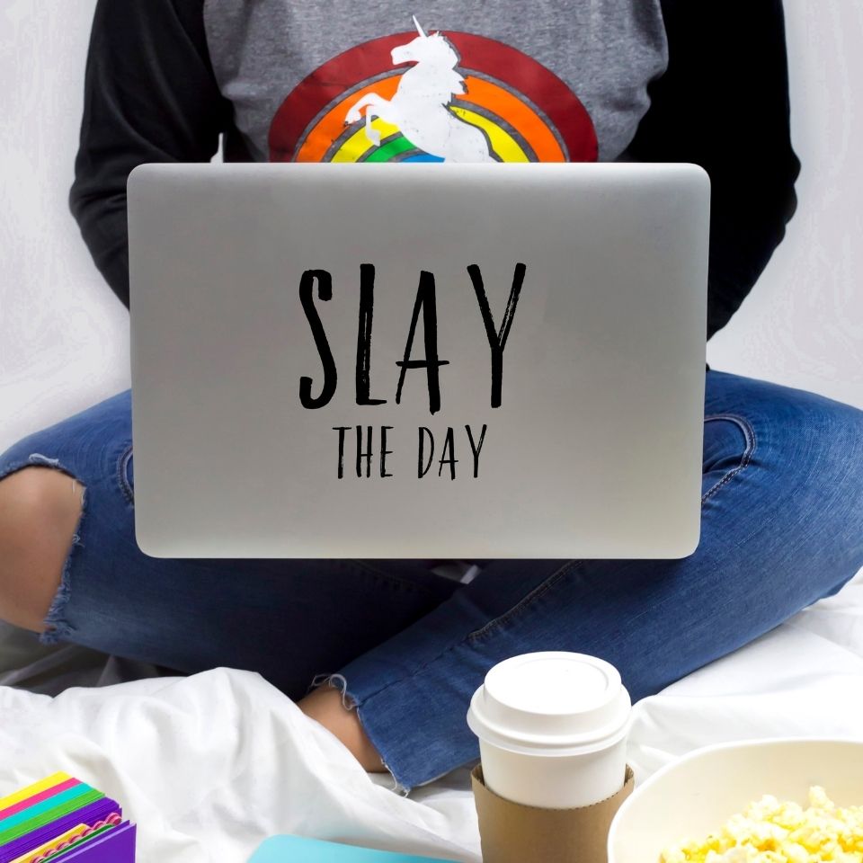 A woman in a rainbow unicorn short working on a laptop with the words "Slay the day" on the back. A cup of coffee is visible by her feet. Her face is not visible.