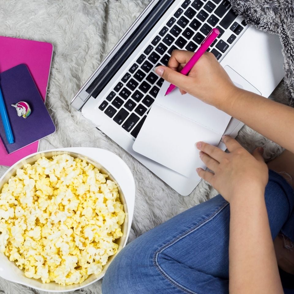 A top down closeup image of a woman writing in a small notebook with a pink pen. She's using her laptop as a table and has a bowl of popcorn. Only her forearms and left knee, wearing jeans, are visible - the rest of her body is not.