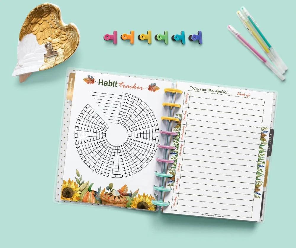 A mockup of an open Happy Planner classic on a light green background showing a habit tracker and a gratitude journal page. There are desk supplies around the planner including colorful binder clips and three gel pens.