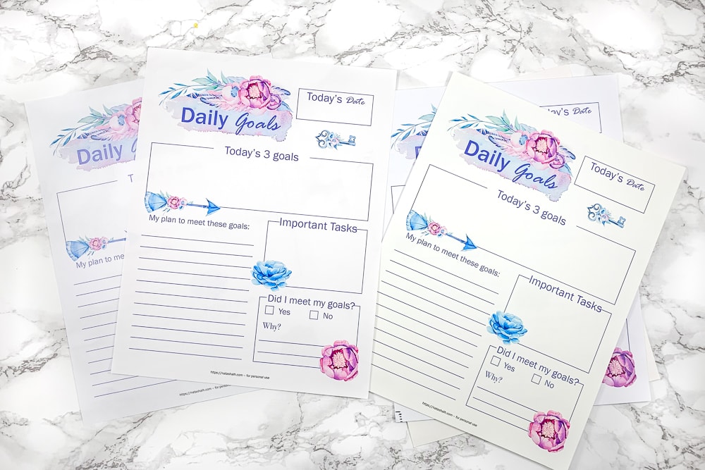 Four copies of the same pink and blue floral daily goals planner page printed on different types of paper. On top are pages printed on presentation paper. Below and to the left a page printed on regular printer paper is visible with much less vibrant colors. The pages are resting on a white faux marble background.