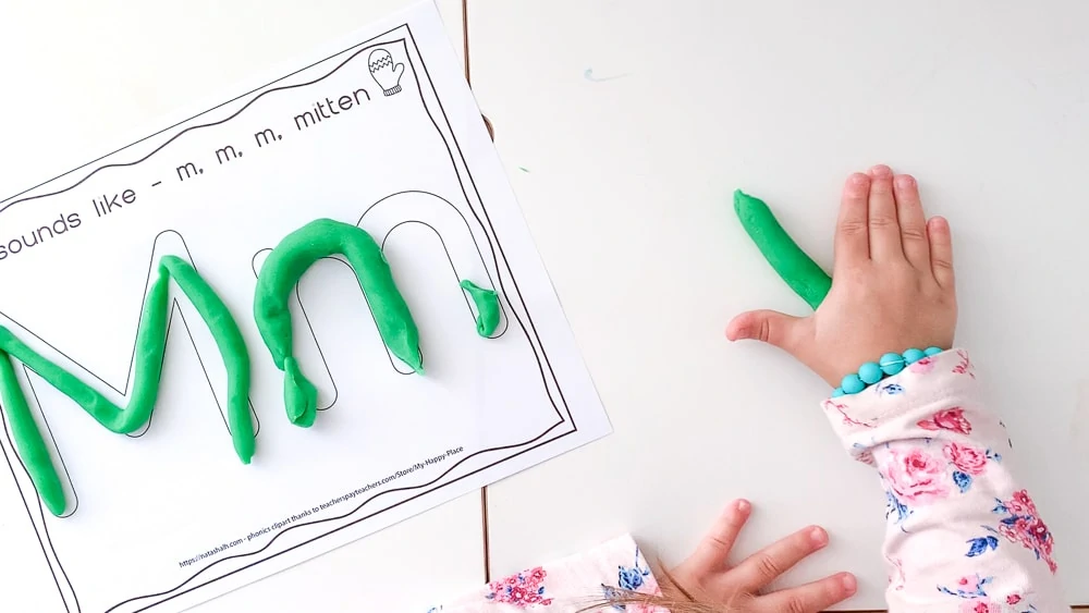 a top down shot of a toddler's hands rolling out green play dough on a white table. Next to her hands is a piece of paper with Mm written in big bubble letters to fill with play dough