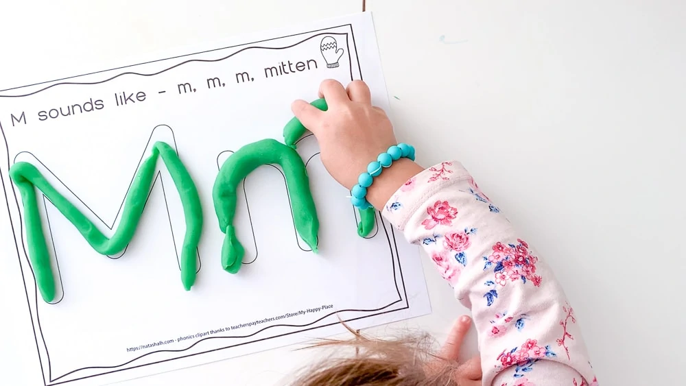 A top down view of a young child with a pink floral shirt on and a blue bracelet placing a piece of green play dough on a letter mat with large bubble letters M and m 