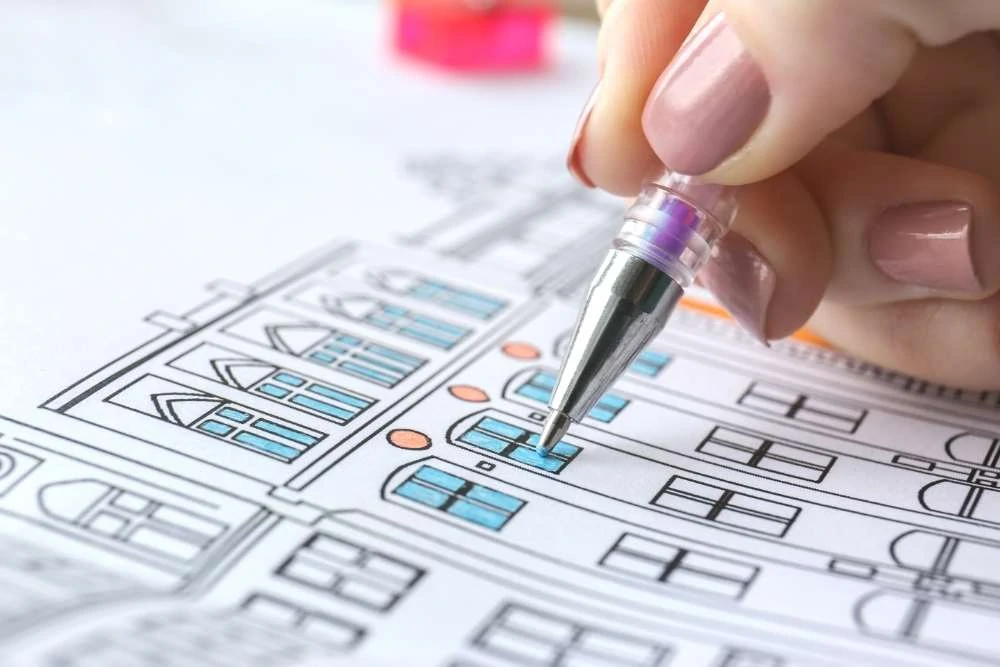 A close up fo a woman's hand using a gel pen to color a window on a drawing of a house bright blue. Two rows of windows are colored blue and there are orange circles above the windows on the lower row.