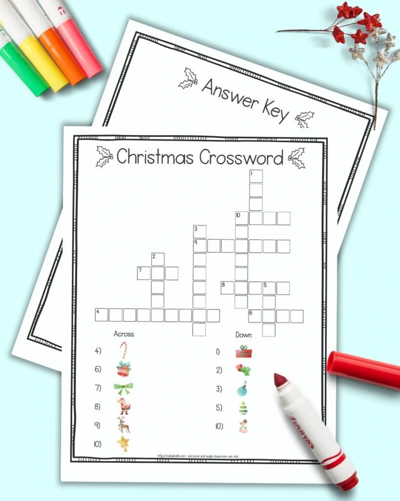 A free printable Christmas crossword puzzle for children featuring image clues instead of text clues. The page has a doodle frame border. The crossword is on a blue background surrounded by children's markers. Behind the page with a crossword puzzle a second page is barely visible. It is the puzzle's answer key.