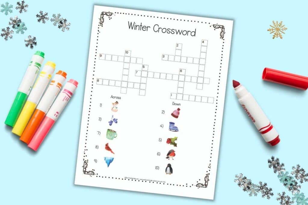 A flatlay of a free printable easy winter crossword with images instead of text-based clues. The page is on a light blue surface with colorful children's markers and snowflake shaped confetti.