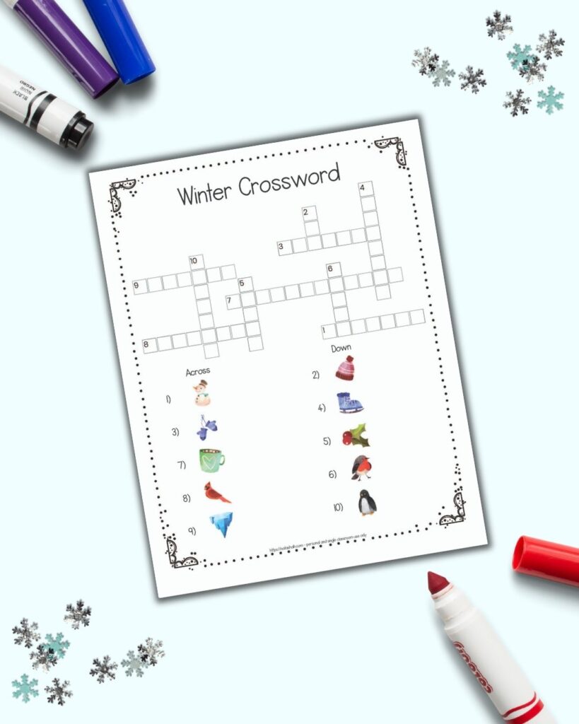 A flatlay of a free printable easy winter crossword puzzle with pictures instead of text clues. The page is on a light blue background with children's markers and snowflake confetti. 