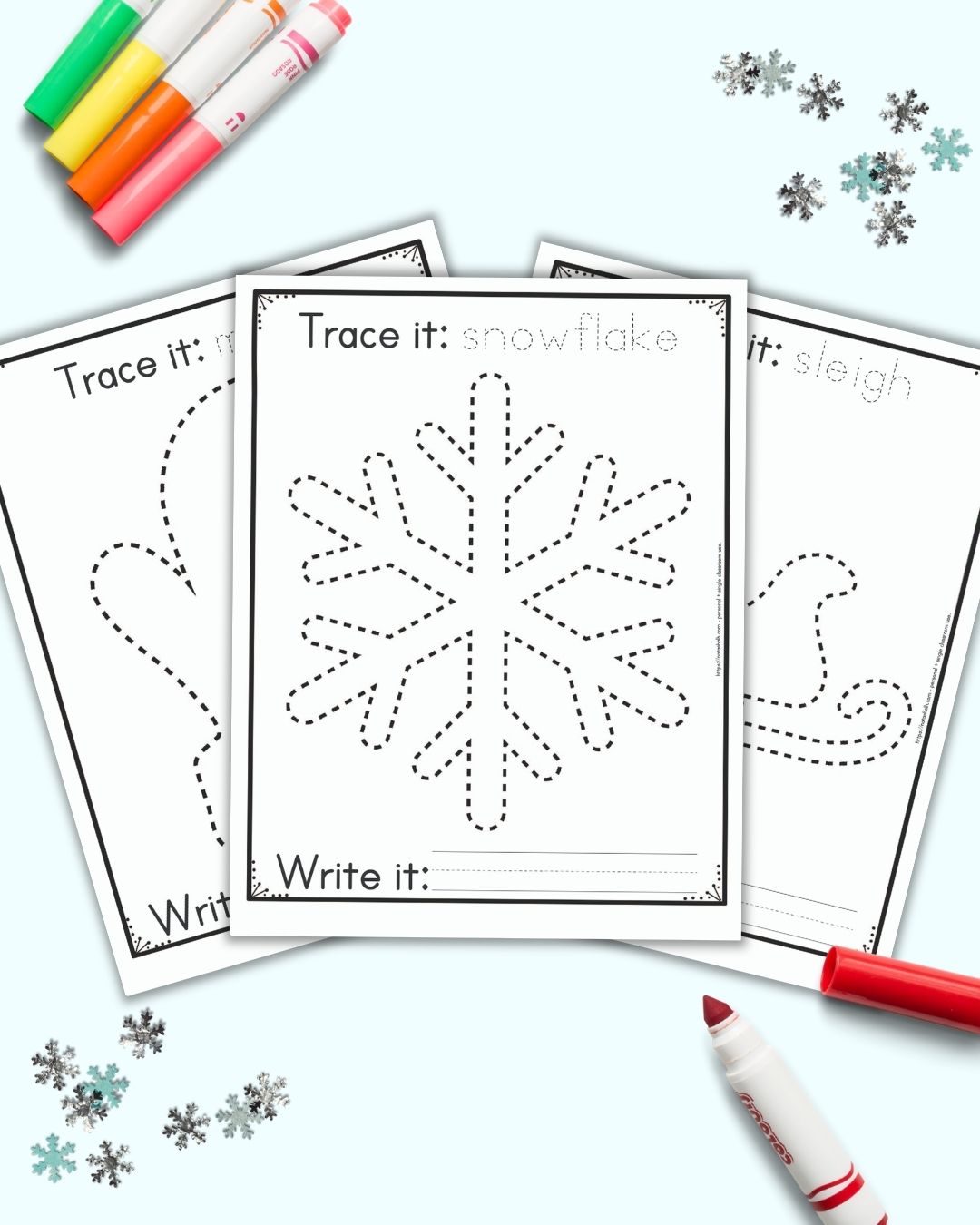 Three free printable winter themed tracing images for preschoolers and kindergarteners. The front and center page has a dotted snowflake to trace. It has the caption "Trace it: snowflake" on top and "write it" with a blank line for writing below the snowflake. Behind the snowflake are a mitten and a sleigh to trace on separate pages. The printables are on a blue background with snowflake confetti and brightly colored children's markers.