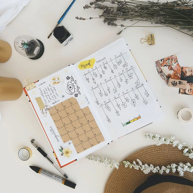 a flatly image showing an open bullet journal with a monthly layout on the left and a mood tracker on the right. The notebook is surrounded y office supplies, a sun hat, lavender, and two gold pillar candles. 