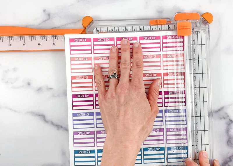 A woman's hand holding a page of printable planer stickers for cutting with a white and orange paper trimer. 
