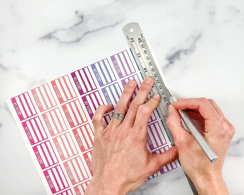 A woman's hands holding a metal ruler and a metal hobby knife against a sheet of printable planner stickers. She is using the hobby knife to cut along the ruler's edge of a row of stickers.