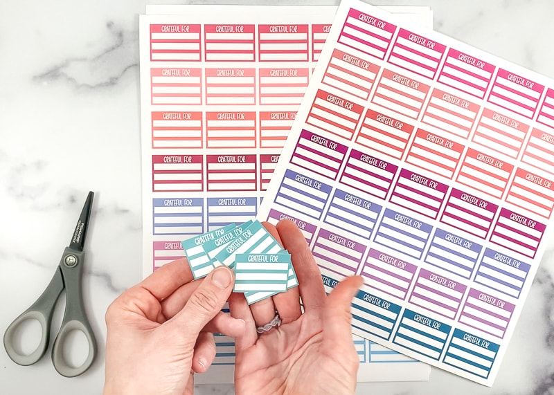 A woman's hands holding 5 small teal box planner stickers. IN the background are two more pages of printed stickers at a small pair of grey scissors. 